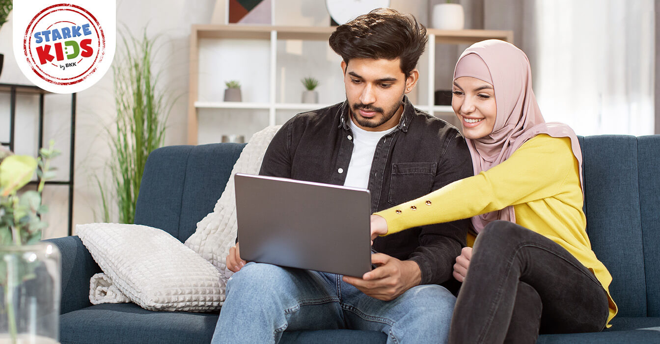 Ein Paar sitzt zusammen auf einem Sofa und betrachtet gemeinsam einen Laptop, die Frau trägt ein gelbes Kopftuch und lehnt sich lächelnd an den Mann.