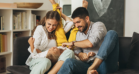 Eine Familie hat Spaß zusammen auf der Couch, mit einem lachenden Kind, das auf dem Rücken liegt, umgeben von seinen fröhlich aussehenden Eltern.