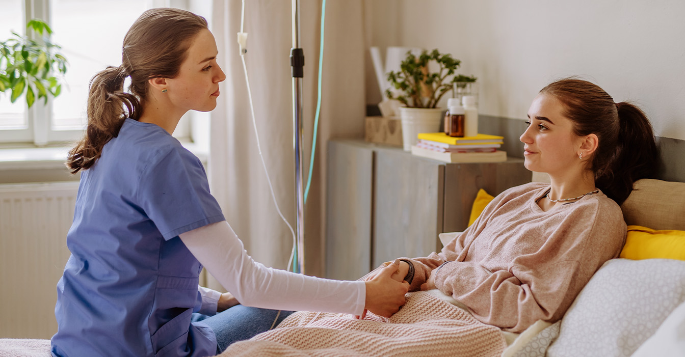 Eine medizinische Fachkraft in blauer Uniform sitzt neben einer lächelnden jungen Person auf einem Sofa; sie hält beruhigend die Hand der Person, die an einen intravenösen Tropf angeschlossen ist.