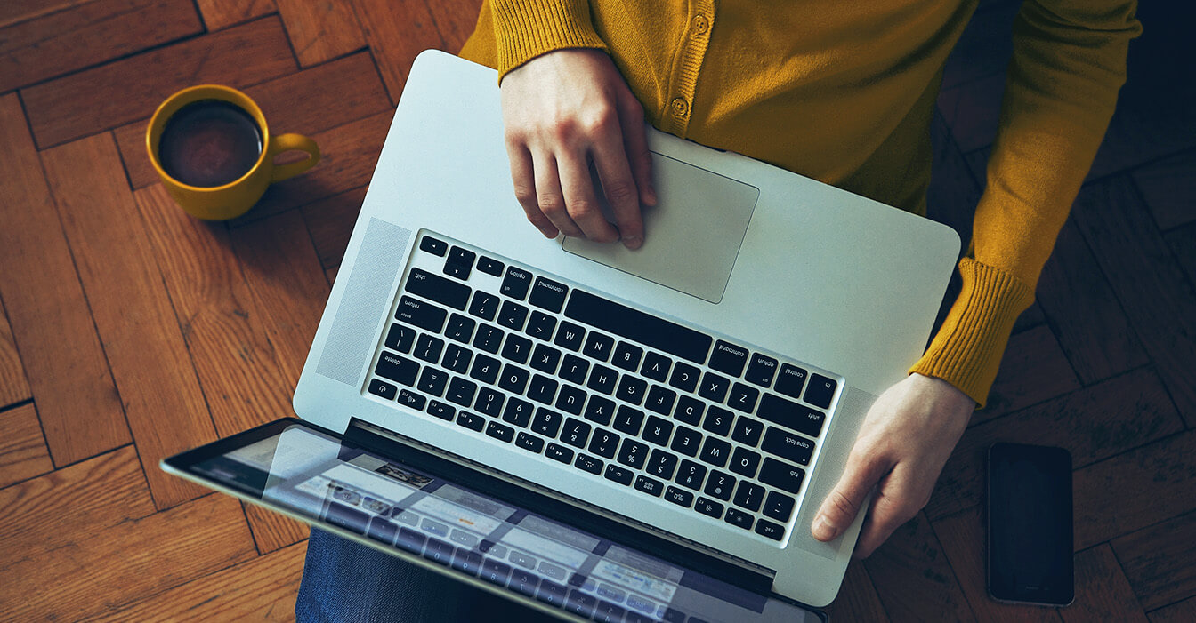 Person bedient ein Laptop auf einem Holztisch, neben ihr eine Kaffeetasse und ein Smartphone.